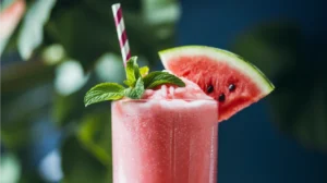 A frosty pink watermelon slushie in a tall glass with fresh mint leaves, a red and white striped straw, and a small watermelon wedge on the rim